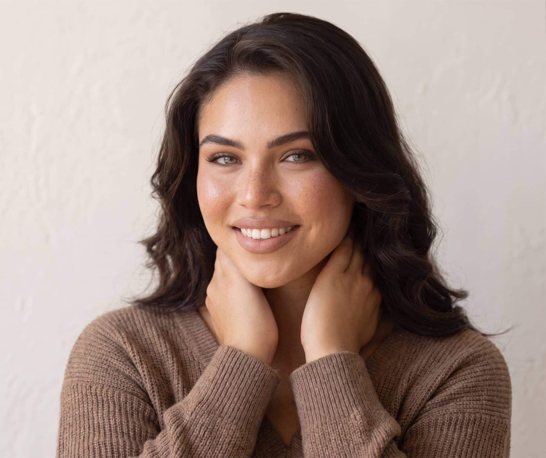 A smiling woman with loose dark wavy hair, hands behind her neck